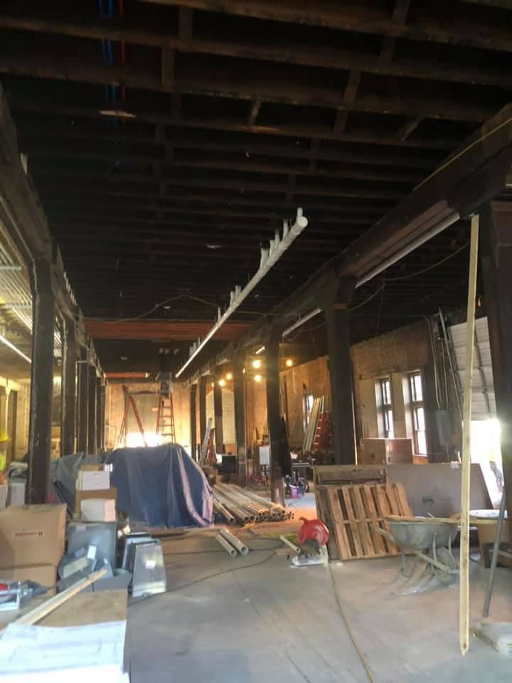 Long, dim construction site with exposed beams, scattered tools, and covered equipment inside a warehouse-like space