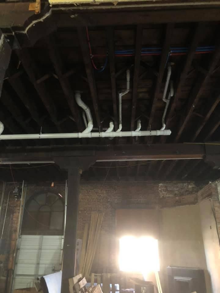 Exposed ceiling beams and pipes in a dim interior, with bright sunlight streaming through a doorway below. Exposed ceiling beams and pipes in a dim interior, with bright sunlight streaming through a doorway below.