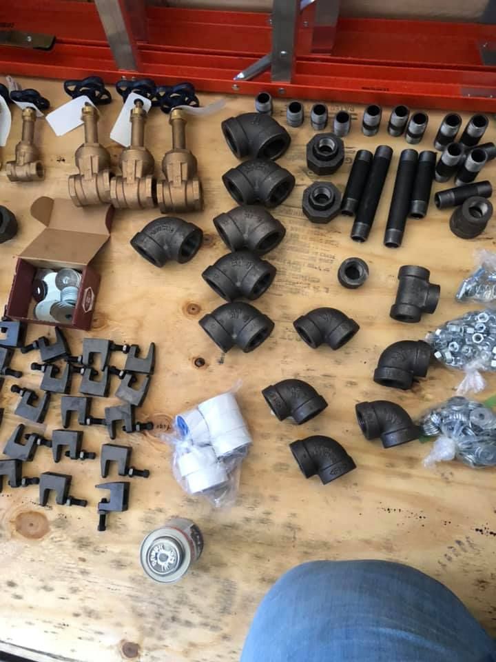 Assorted black and metal pipe fittings, clamps, and hardware pieces arranged on a wooden workbench. Assorted black and metal pipe fittings, clamps, and hardware pieces arranged on a wooden workbench.