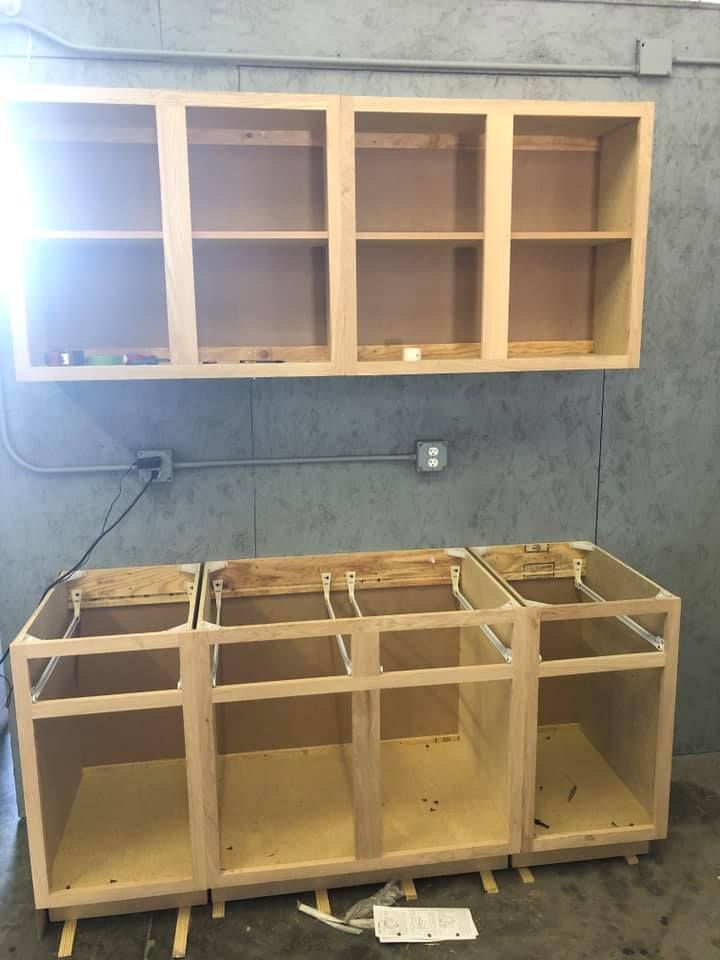 Unfinished wooden kitchen cabinets installed against a gray wall, with exposed wiring and tools below. Unfinished wooden kitchen cabinets installed against a gray wall, with exposed wiring and tools below.