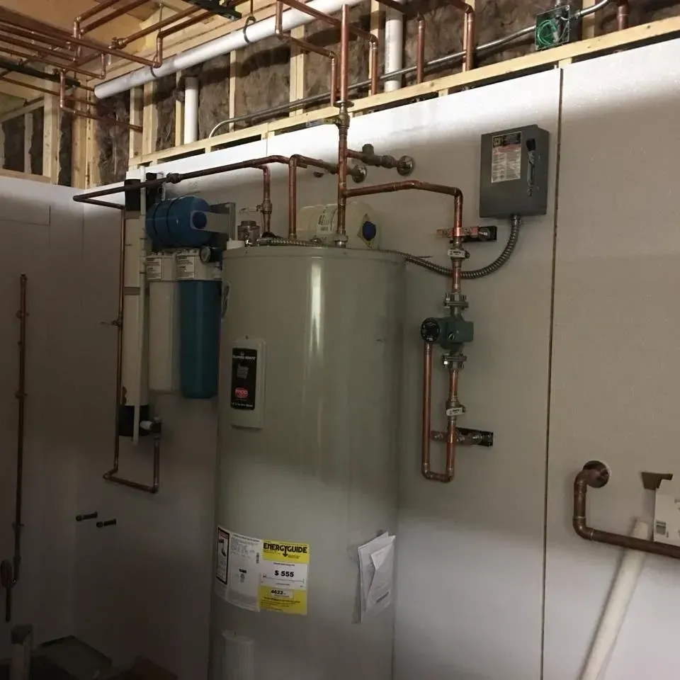 Utility room with a large white water heater, blue filter tank, and exposed pipes on the wall.
