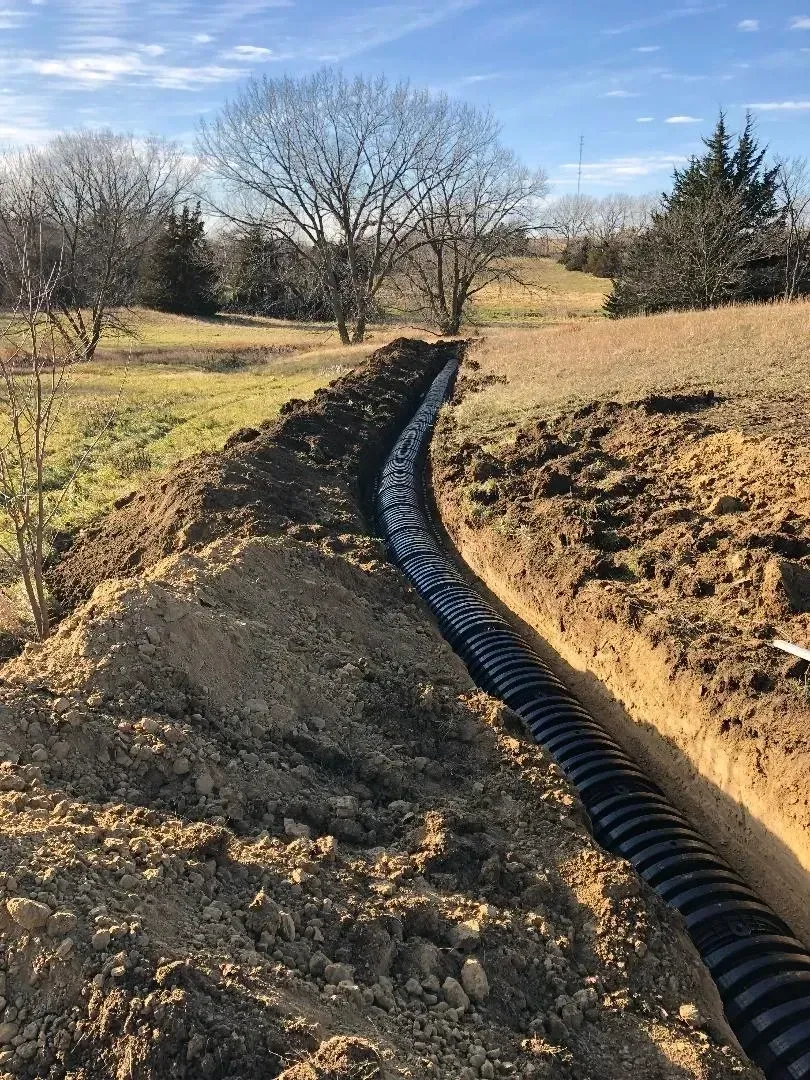 Black drainage pipe runs through a dirt trench across a grassy field with leafless trees and a clear sky.
