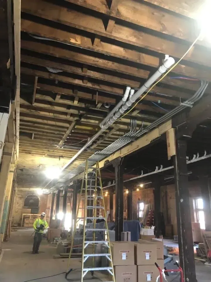 Interior construction site with ladder, stacked boxes, workers, and exposed wooden beams over a partially built ceiling Interior construction site with ladder, stacked boxes, workers, and exposed wooden beams over a partially built ceiling