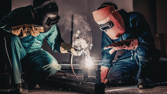 Two welders in protective gear working on a bright welding seam in a dark workshop.