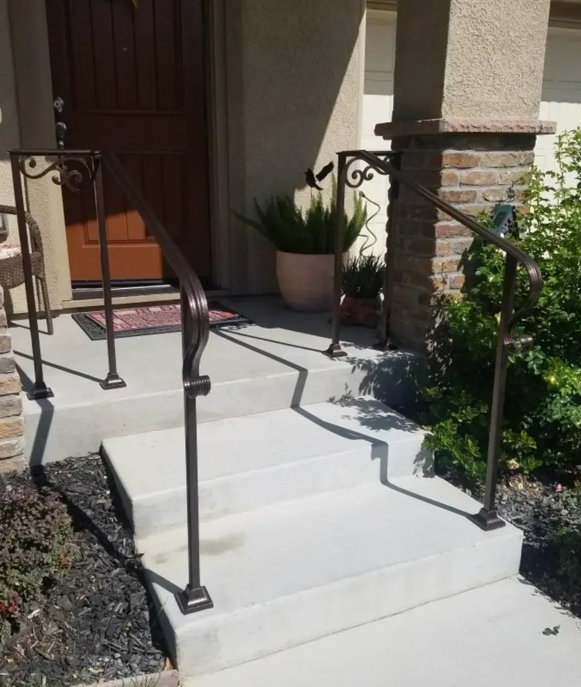 White front steps with black railings leading to a brown entry door, beside a stucco porch and potted plant