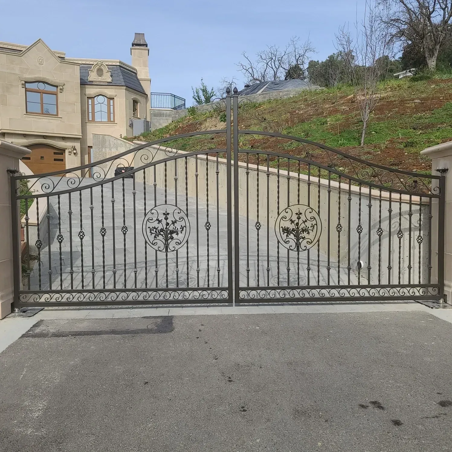 Black wrought-iron driveway gate beside a cream house on a sloped hillside under a clear sky
