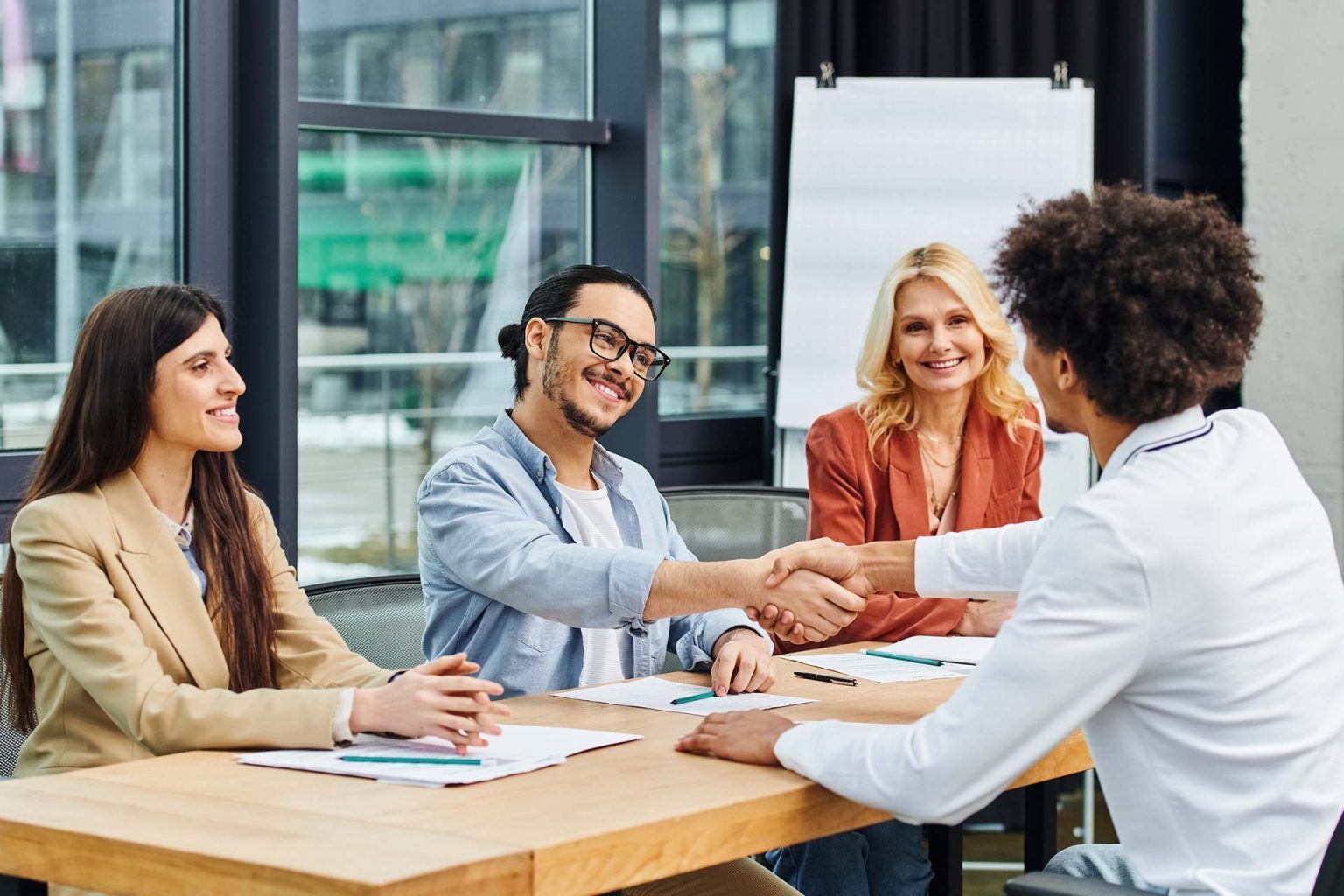 Four people in a meeting room shaking hands around a table, with papers and a flip chart nearby.