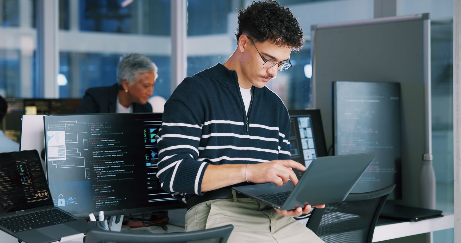 Person using a laptop in a tech workspace with code on monitors in the background.