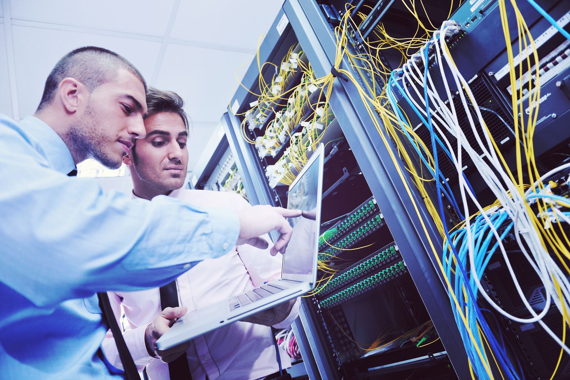 Two technicians inspecting server racks, pointing at laptop amid network cables.