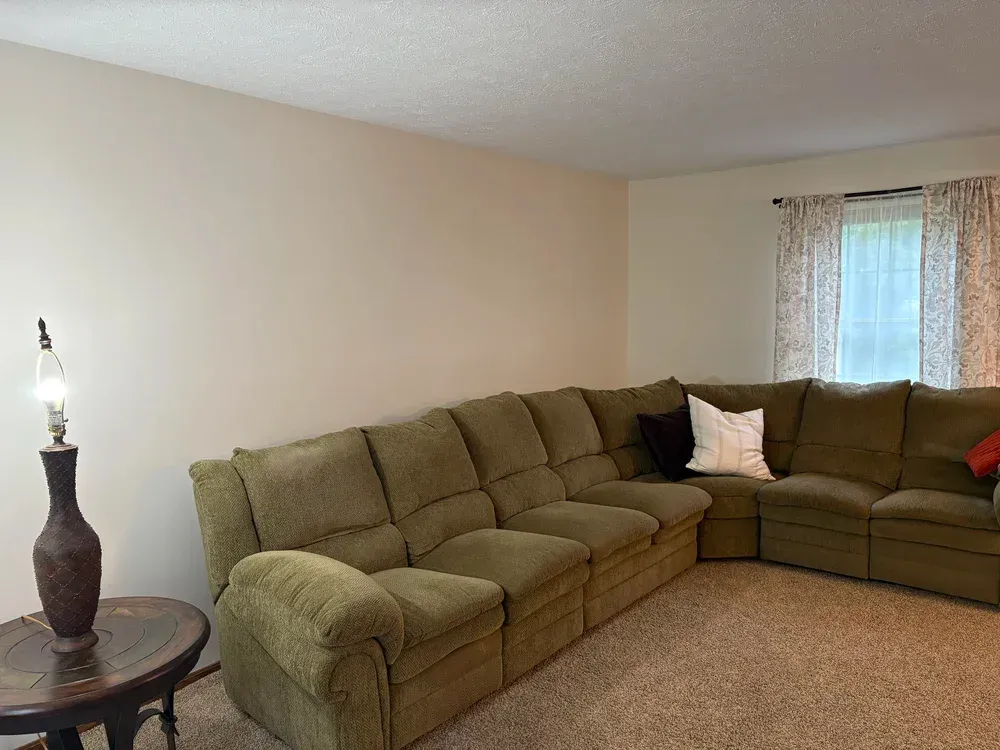 A living room with an olive green sectional sofa, a lamp on a table, and a window with floral curtains.