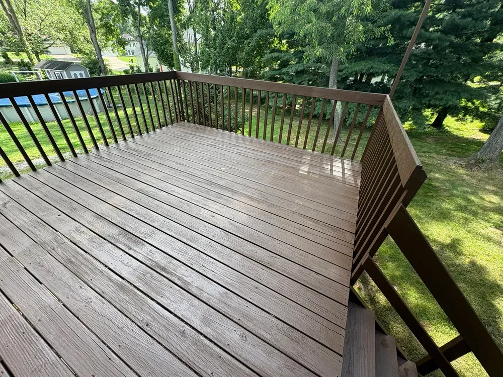 Brown wooden deck with railings, viewed from above, surrounded by trees and grass.