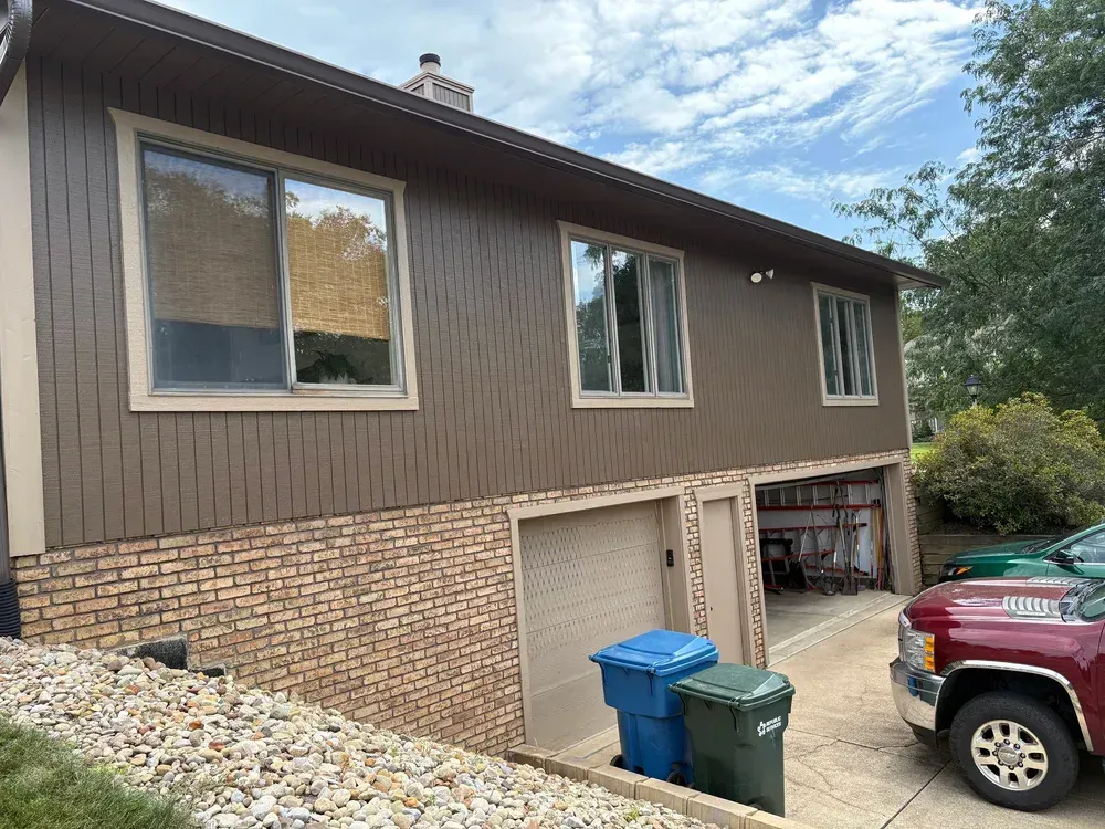 Brown-sided house with brick base, two garage doors, and windows; a truck and trash bins are in front.