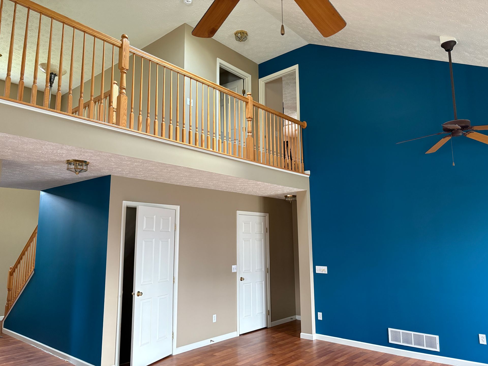 Interior view of a house with a loft, featuring blue and beige walls, wooden staircase and flooring, and ceiling fans.