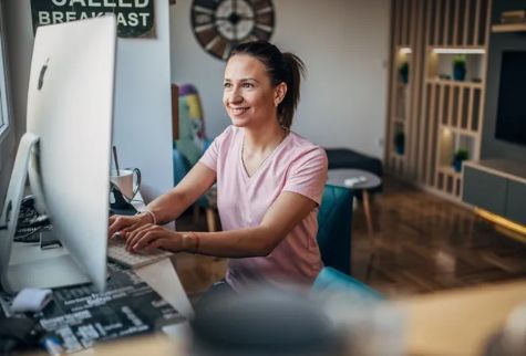 Woman smiling while working on a computer at a desk in a well-lit living room.