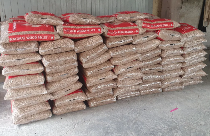 Sacchi di pellet di legno accatastati contro un muro. I sacchi sono color marrone chiaro con scritte rosse, in un ambiente da magazzino.