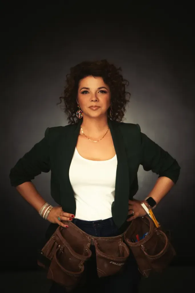 Woman standing with hands on hips, wearing a black jacket, white top, and brown tool belt against a dark background