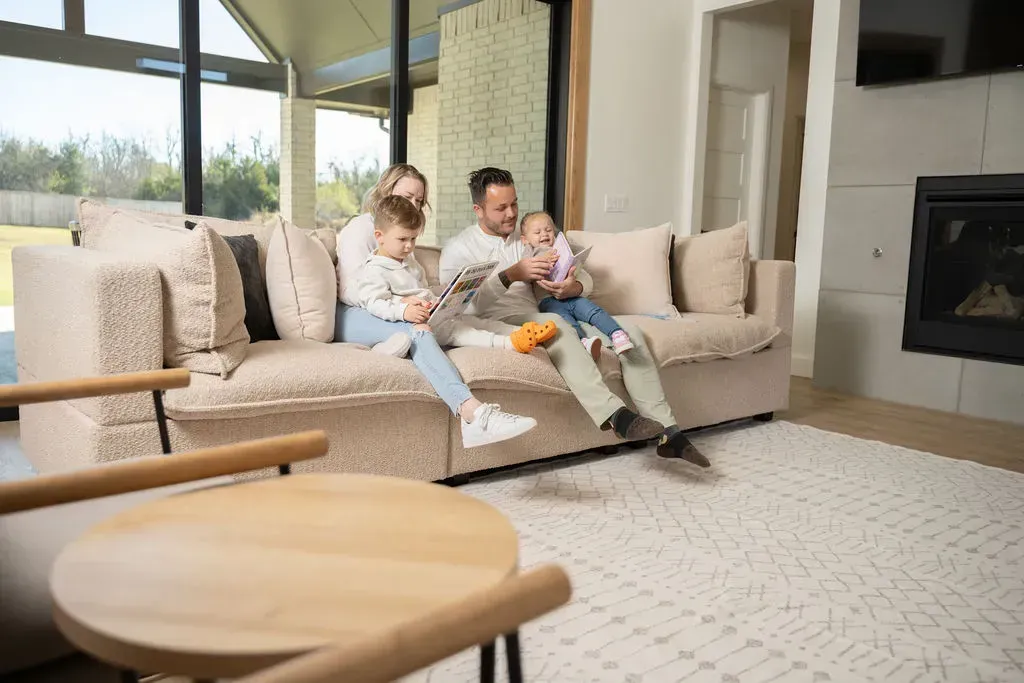 Family sitting on a beige sofa in a bright living room, holding a baby near a fireplace.