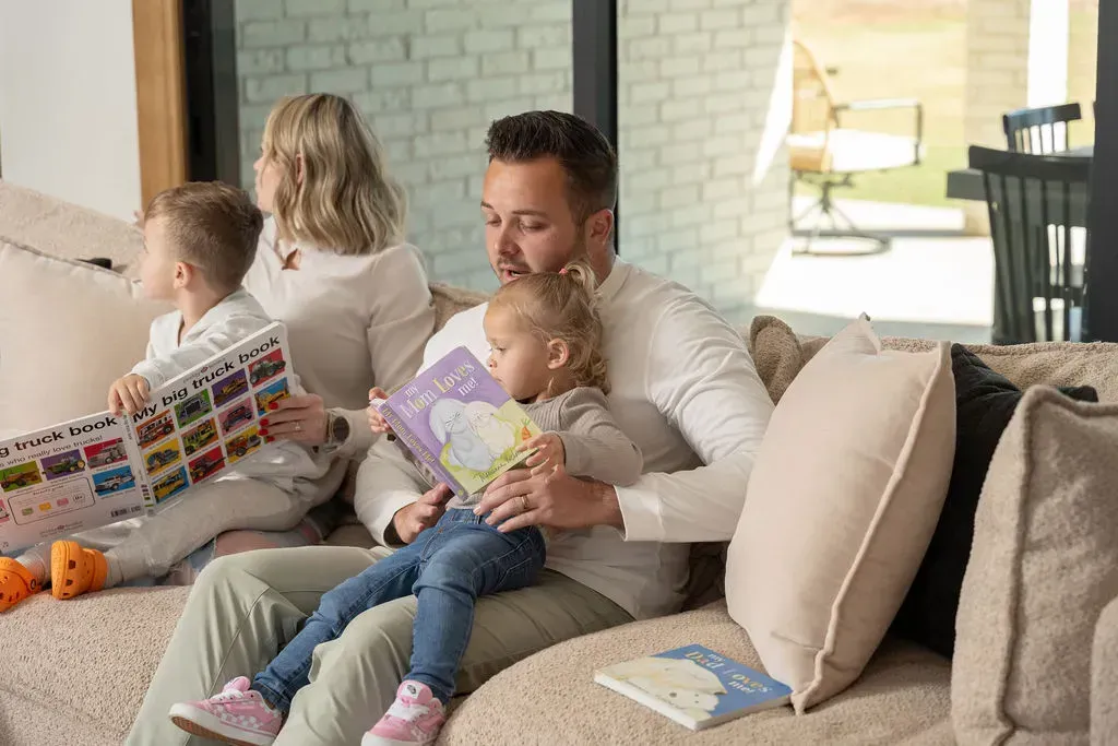 Family seated on a beige sofa, reading books together in a bright living room.