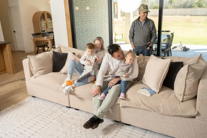 Family sitting on a beige couch in a bright living room, with a man standing behind them.