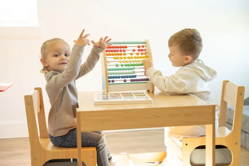 Two toddlers playing with a colorful abacus at a small wooden table in a bright room