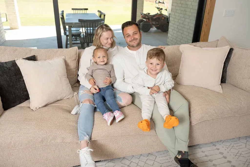 Family of three sitting on a beige sofa in a bright living room, smiling at the camera.