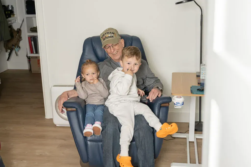 Three people sitting together on a blue chair in a bright living room, one holding a child on each side