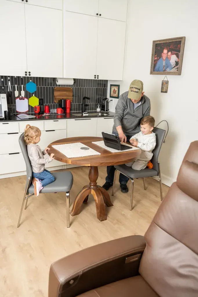 Three people around a dining table in a bright kitchen, with a standing adult and two seated children.