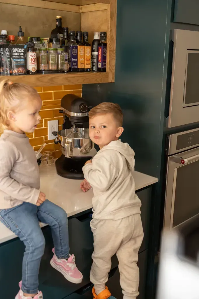 Two children in a kitchen beside a stand mixer, one on the counter and one standing nearby.