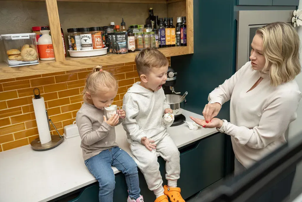 Two children sitting on a kitchen counter while an adult washes one child's hands at the sink.