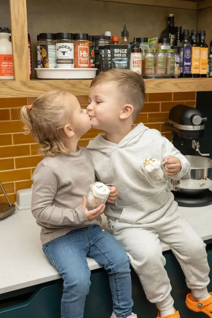 Two children sitting on a kitchen counter, kissing and holding ice cream near a coffee machine and bottles.