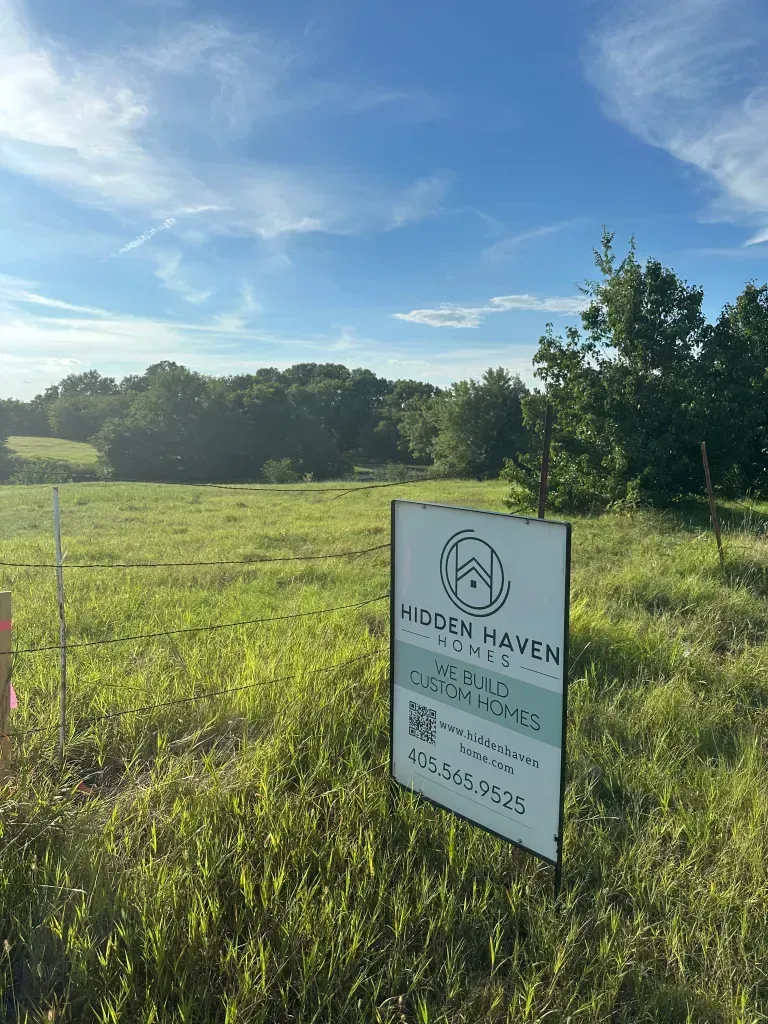 Real estate sign in a grassy field with trees and blue sky behind it.