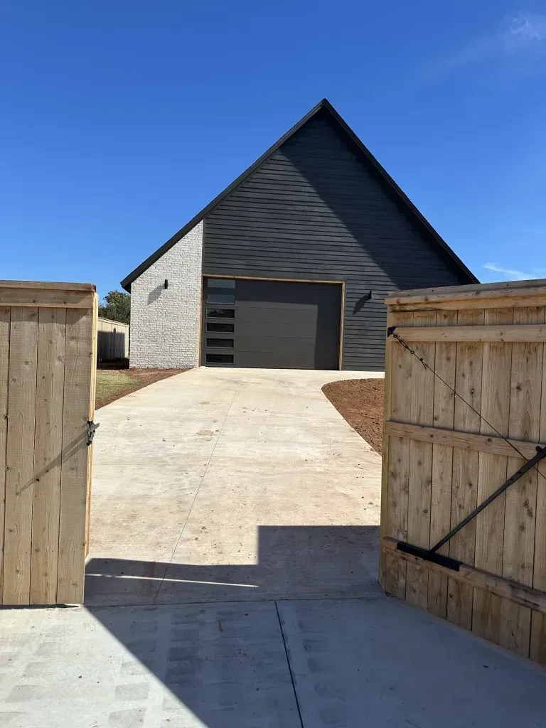 Open wooden gates leading to a driveway and dark gray garage under a clear blue sky