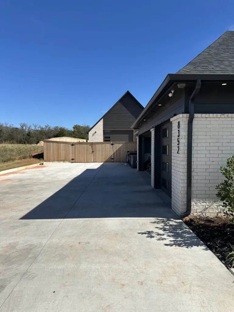 Driveway beside a white brick house under a clear blue sky, leading to a fenced backyard.