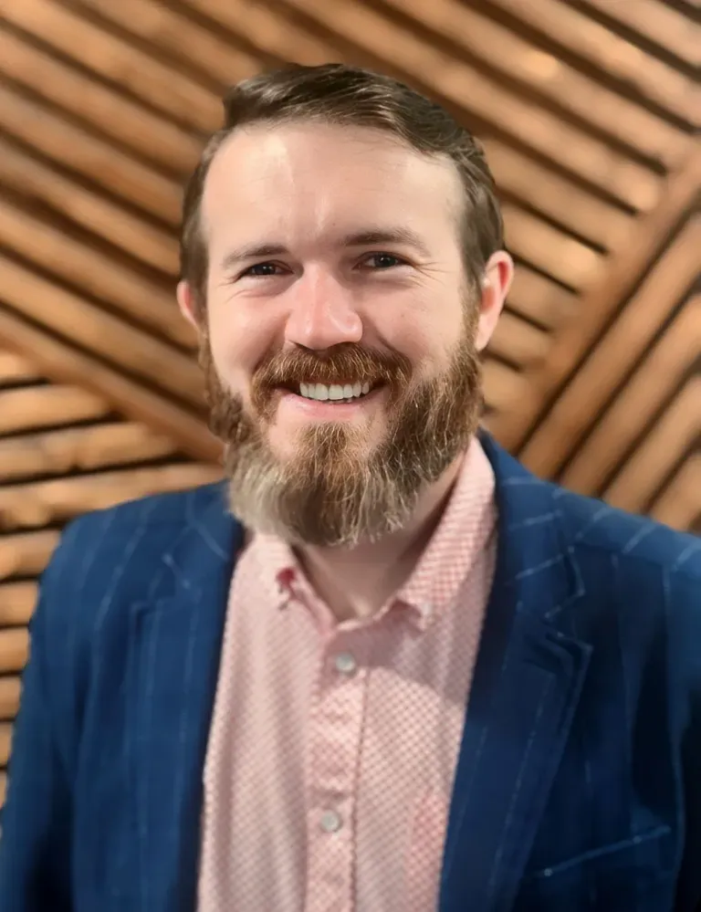 Smiling bearded man in a blue blazer and pink shirt against a wooden slat background