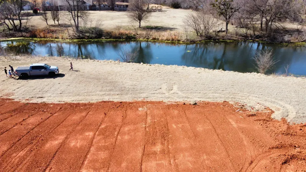 Car parked beside a calm lake, with red dirt foreground and dry hills in the background