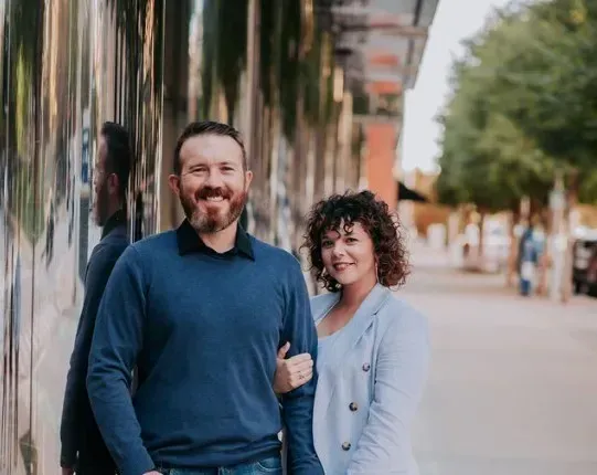 Two people smiling on a palm-lined sidewalk, one in a blue sweater and the other in a light jacket.