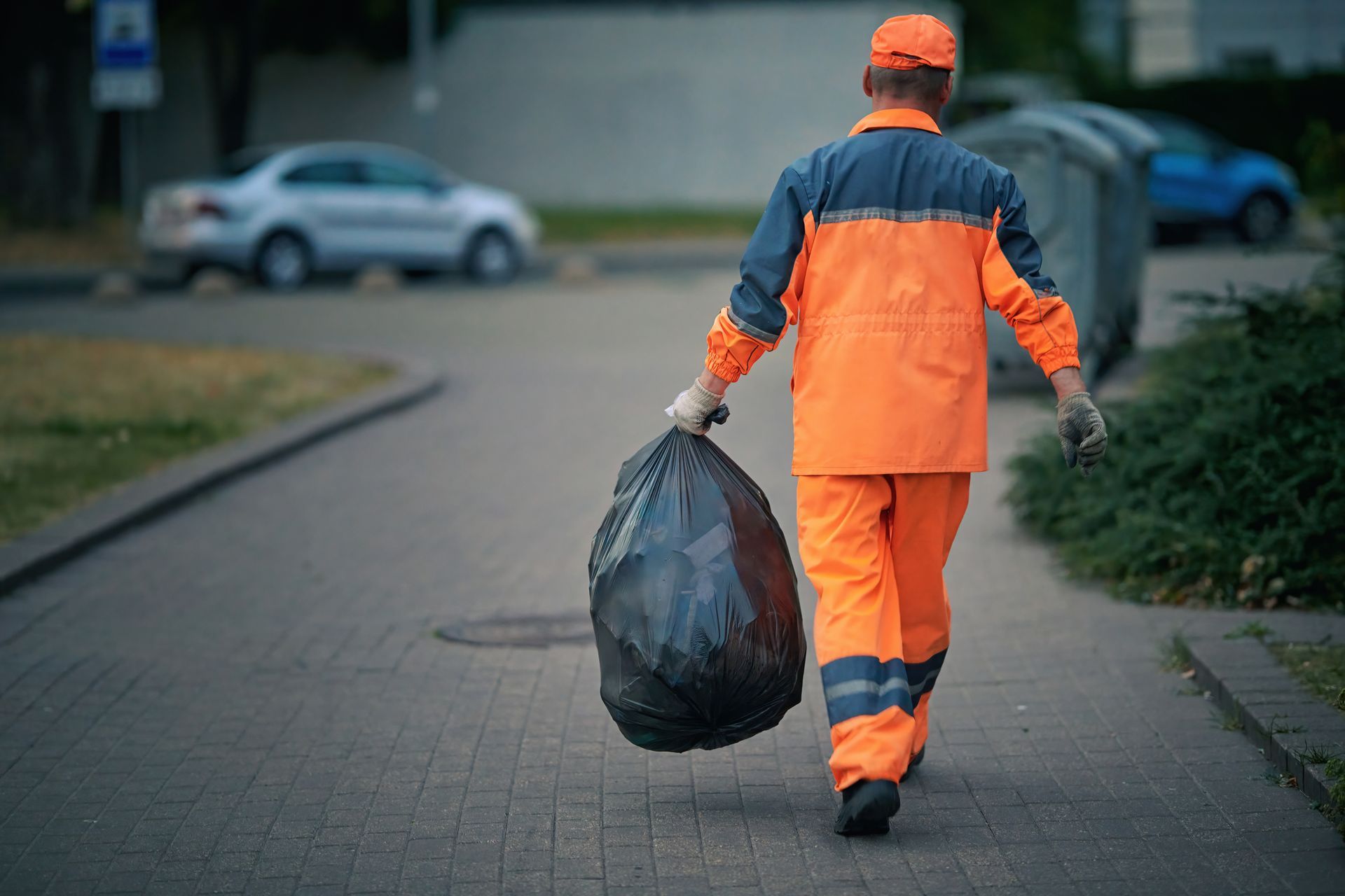 A garbage man is walking down a sidewalk carrying a large black trash bag.