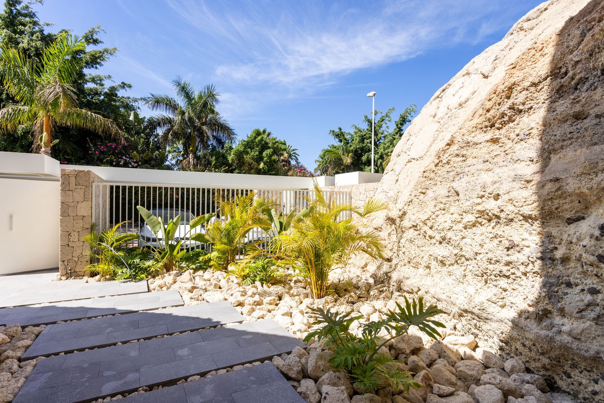 Sunny courtyard with stone path, tropical plants, white fence, and a large rock under a blue sky