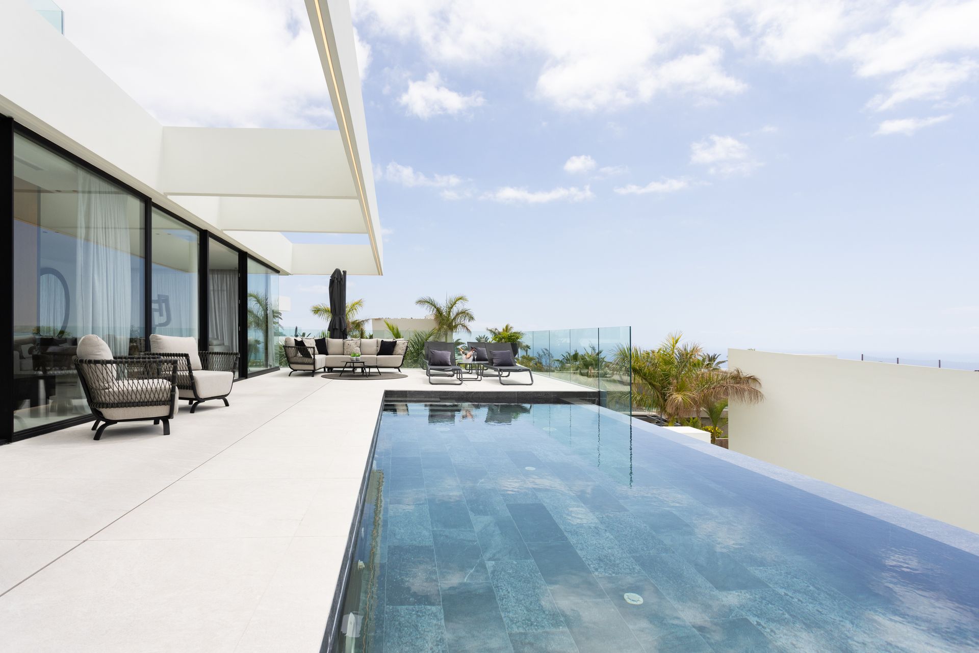 Modern patio with glass doors, lounge chairs, and an infinity pool overlooking the ocean