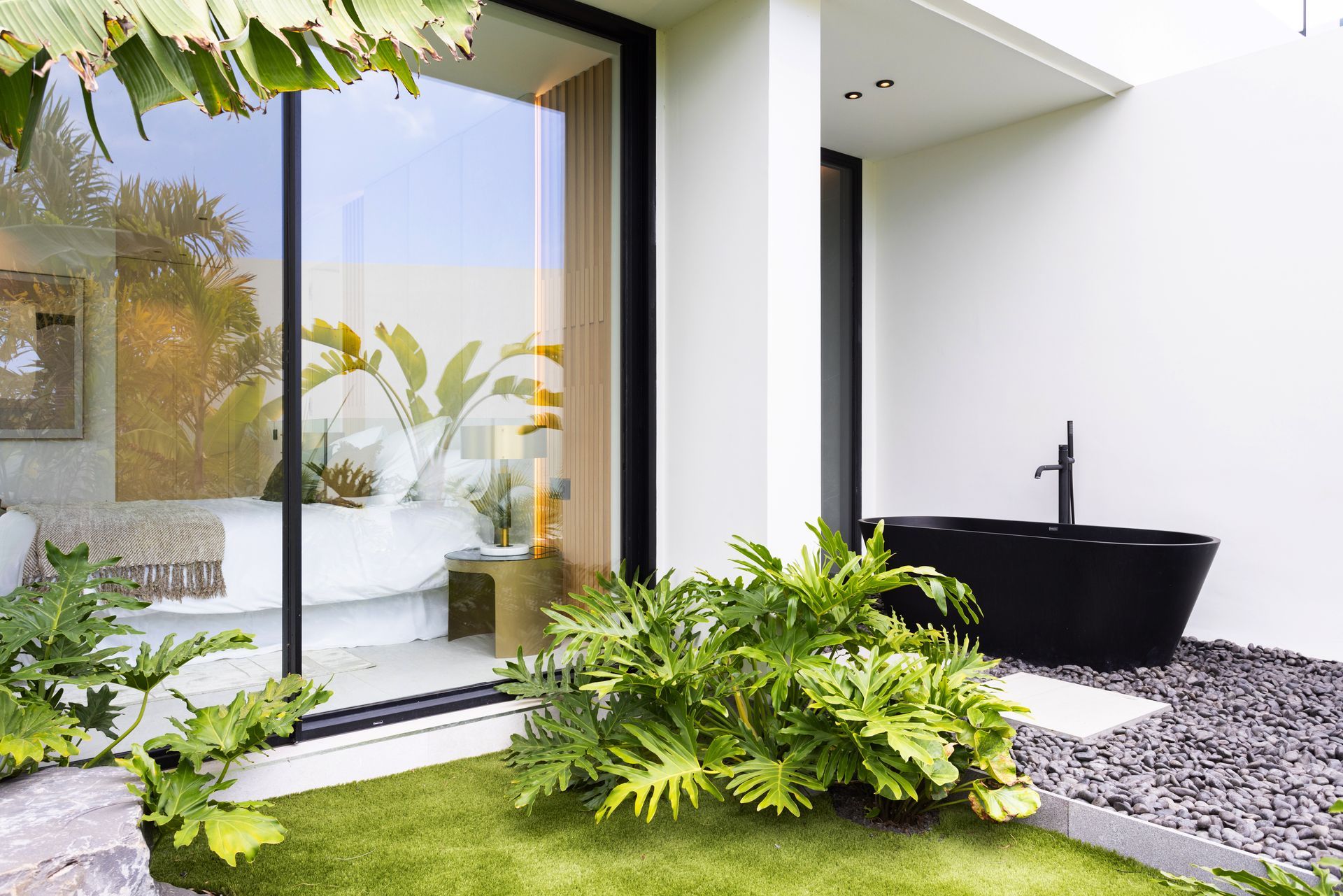 Modern patio with sliding glass door, black bathtub, and lush green plants beside a pebble path