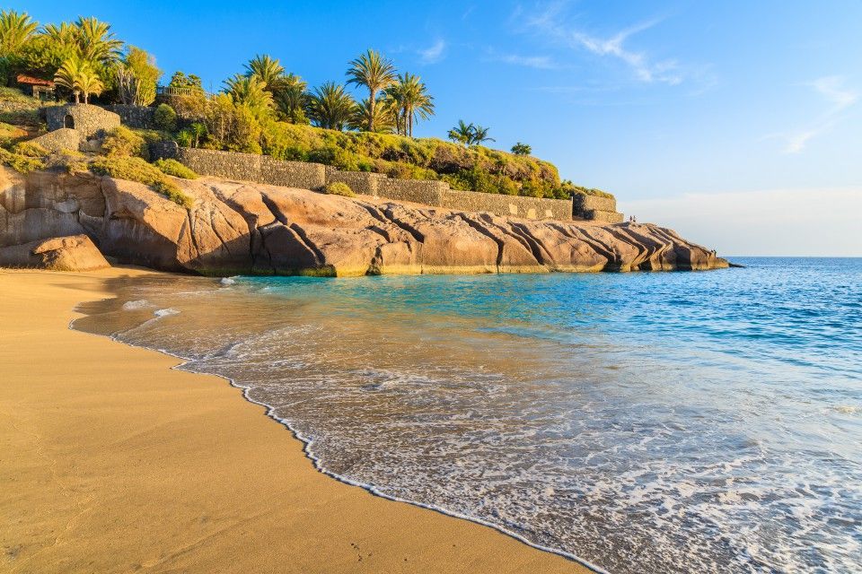 Sandy beach with gentle waves, rocky cliff, and palm trees under a clear blue sky