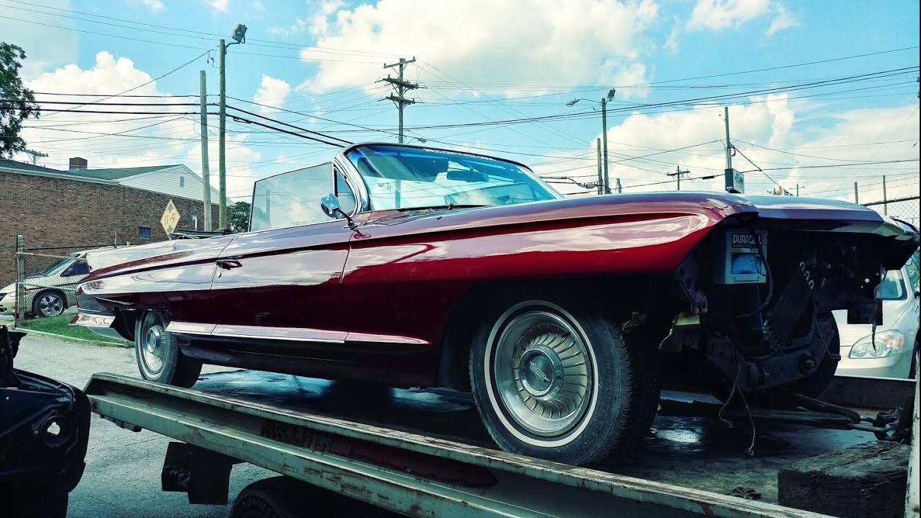 Red convertible car on a flatbed tow truck. The car has missing front parts, under a cloudy sky.