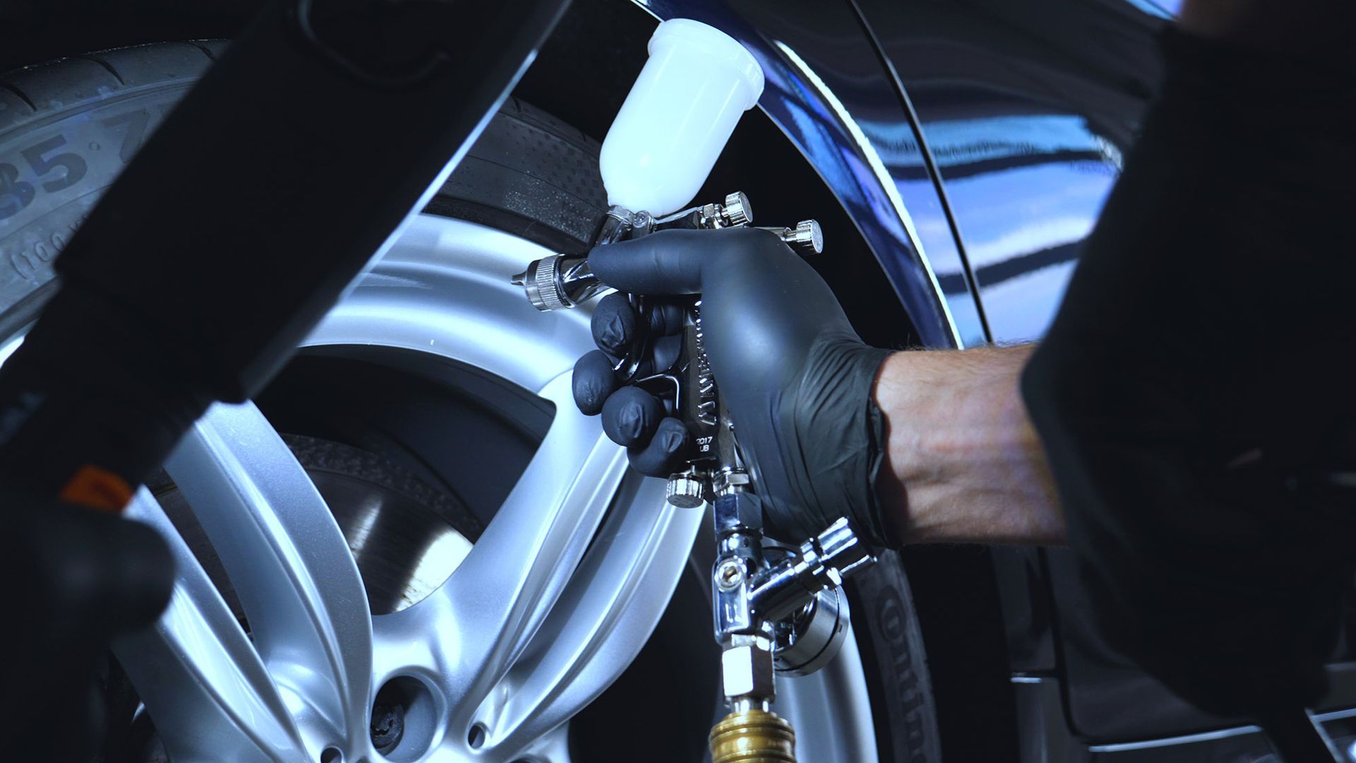 Person in black gloves using a paint sprayer on a car wheel in a garage.