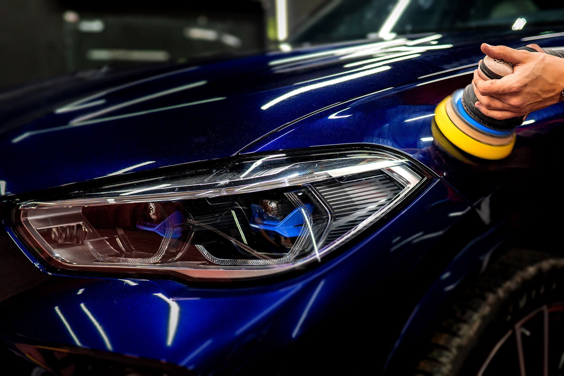 A person polishes a dark blue car with a rotary buffer, focusing on the headlight.