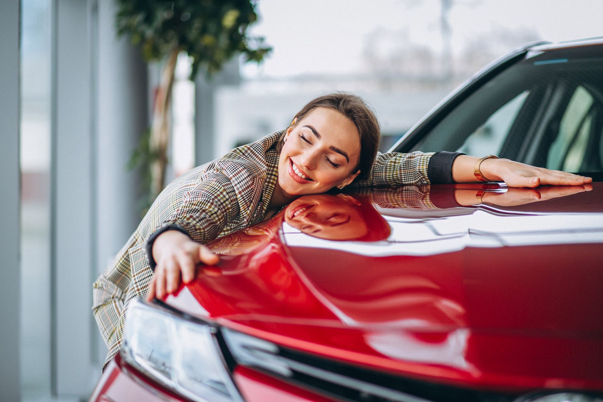 Woman embraces red car in a showroom, smiling with eyes closed.