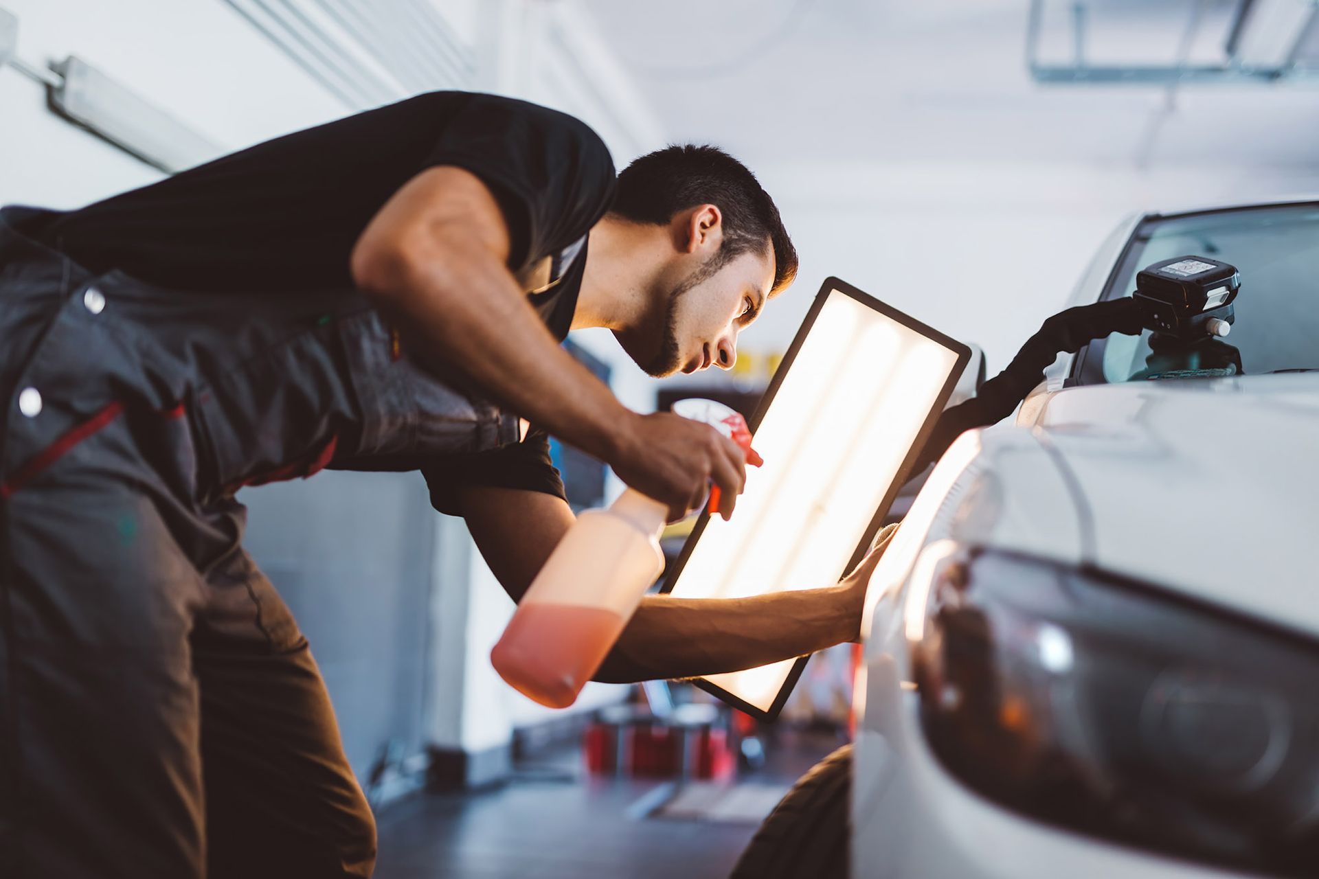 Mechanic inspecting car with light, spraying liquid. Garage setting.