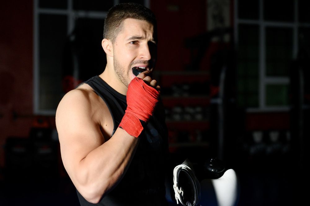 Boxer with red hand wraps, biting his mouthguard, inside a dark gym.