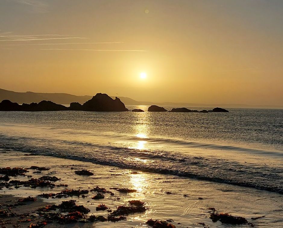 Sunset over ocean with golden light reflecting on the water and beach; rocky outcroppings.