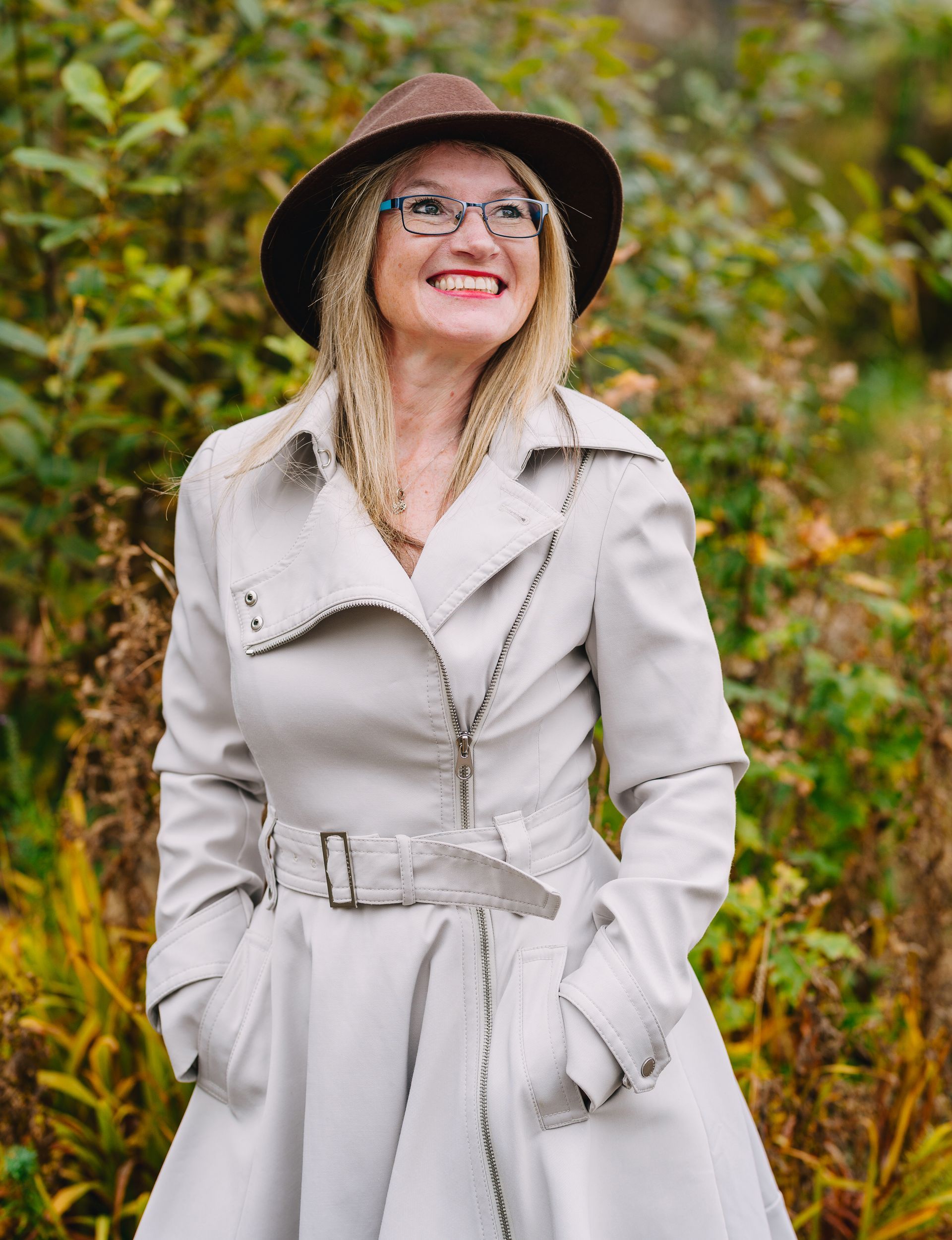 Woman in a light trench coat and hat smiling outdoors among autumn foliage.