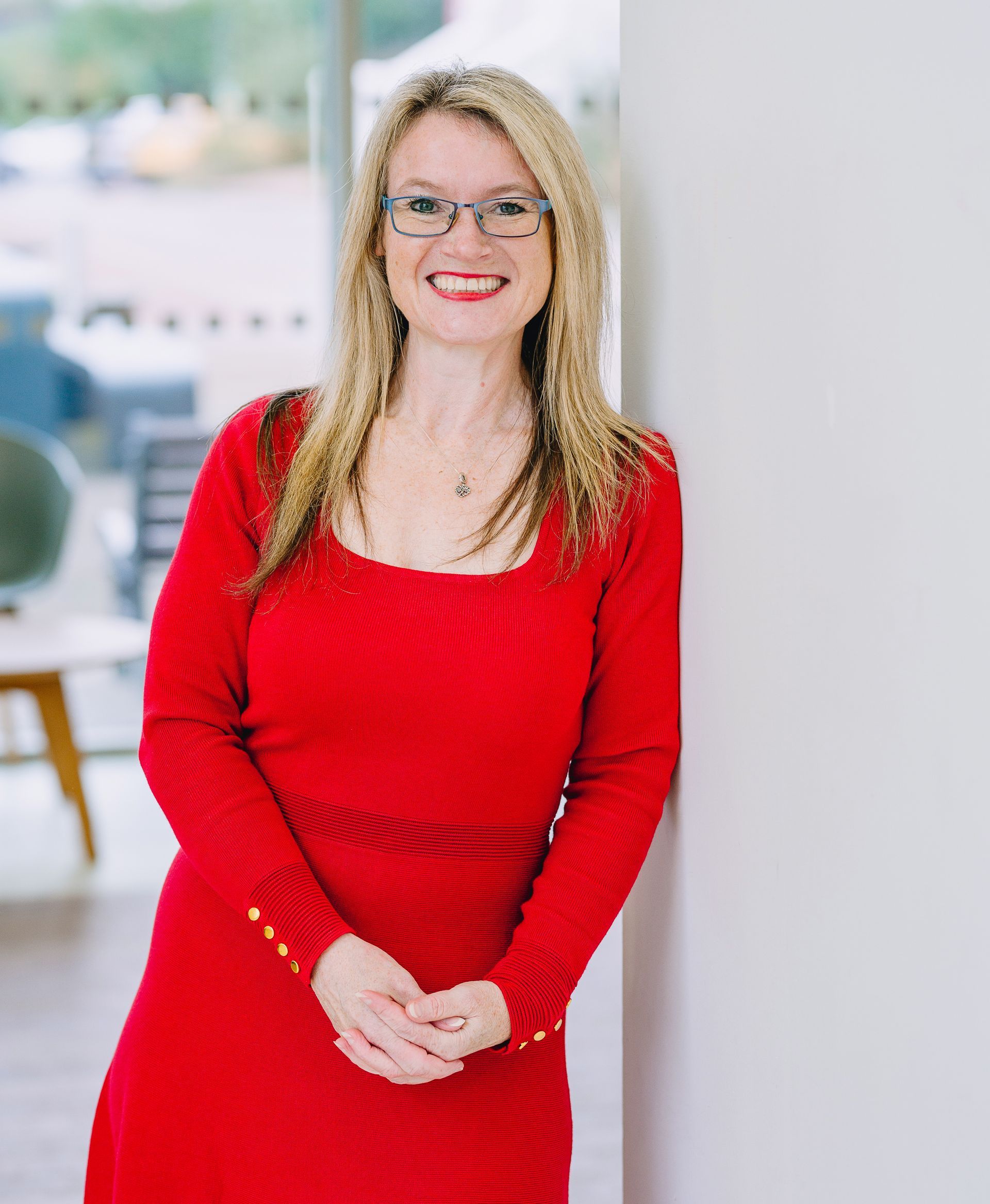 Woman in a red dress and glasses standing indoors, smiling with hands clasped.